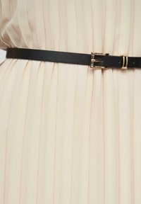 Une ceinture en cuir noir avec une boucle en ton doré, serrée sur un tissu plissé beige clair avec de subtiles rayures roses.