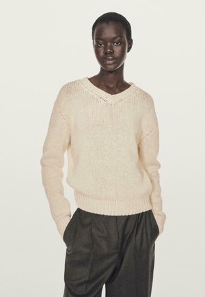 Lyse beige strikket sweater med V-hals, ribbet kant og lange ærmer, parret med mørkegrå plisserede bukser.