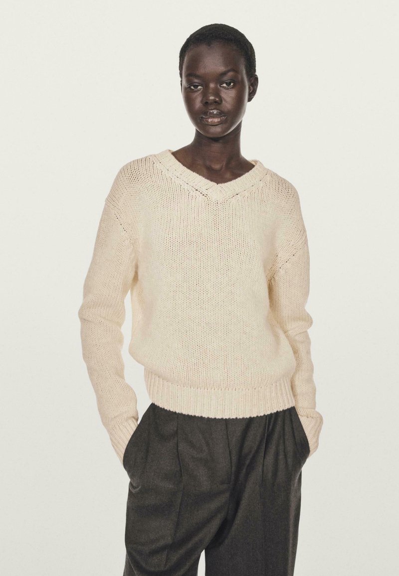 Pull en maille beige clair avec un col en V, ourlet côtelé et manches longues, associé à un pantalon plissé gris foncé.