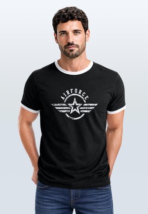 Mann mit kurzen lockigen Haaren und Bart trägt schwarzes T-Shirt mit weißem Rand und vintage weißem Airforce-Stern-Logo, steht mit Händen in den Taschen.