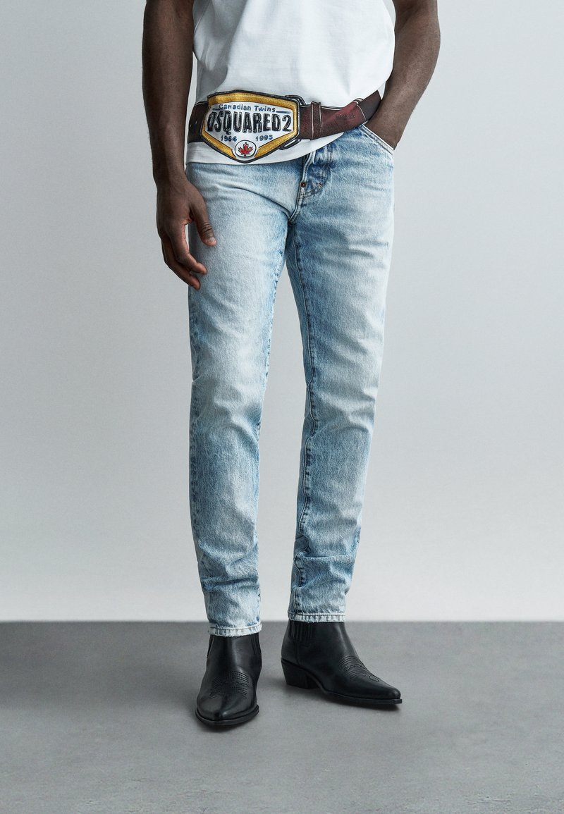 Homme portant un jean en denim bleu clair, des bottes noires à bout pointu, une chemise blanche, et une grande boucle de ceinture avec le texte « DSQUARED2 » et un drapeau canadien.