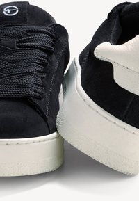 Černé semišové tenisky s bílou gumovou podrážkou, s texturovaným povrchem, plochými shoelaces a logem na jazyku.
