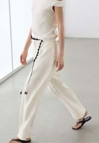 Person walking indoors wearing cream wide-leg pants with a black beaded belt, cream short-sleeve top, and black flip-flop sandals.