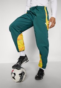 Pantaloni sportivi turchesi con accenti laterali gialli e arancioni; materiale leggero. Scarpe da calcio nere appoggiate su un pallone da calcio decorato.