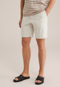 Pantaloni scurți din in de culoare bej deschis, cu dungi verticale albe, având buzunare laterale; purtați cu sandale negre din material țesut.