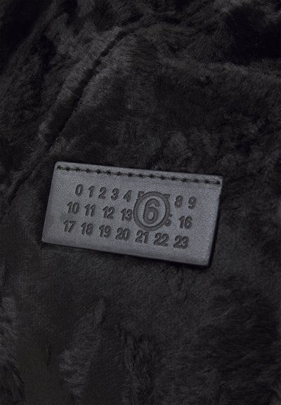 Têxtil de pelúcia sintética preta com uma textura suave e macia, apresentando uma etiqueta retangular de couro preto com números em relevo e um logotipo circular.