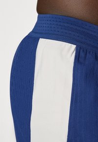 Blå atletiske shorts med en tekstureret talje, der har hvide sidedele og en glat stoffinish. Synlig syning.