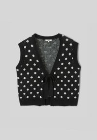 Veste noire sans manches en tricot avec des pois blancs, col en V profond et fermeture à nouer devant, sur un fond gris uni.