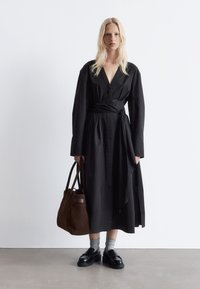 Schwarz langärmliges Kleid mit V-Ausschnitt und Taillengurt, aus glattem Stoff. Kombiniert mit einer braunen Wildleder-Tote und schwarzen Plattformslippers.