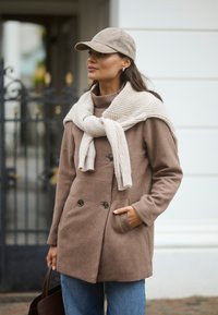 Cappotto doppiopetto in misto lana marrone con grandi bottoni neri, abbinato a una sciarpa in maglia a coste color crema e a un berretto grigio. Indossato con jeans di denim.