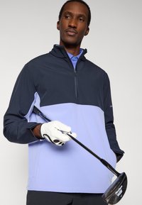 Pulover de golf în nuanțe de albastru închis și lavandă deschis, cu guler cu fermoar și mâneci lungi. Purtat cu o mănușă de golf, ținând un crosă. Textură netedă.