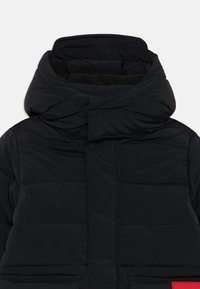 Doudoune noire avec un col haut, texture matelassée, fermeture à boutons sur le devant et une étiquette en tissu rouge sur le côté gauche.