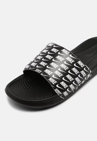 Black slide sandal with a textured sole, featuring a wide strap adorned with repeated white "Nike" logos. Smooth synthetic material.