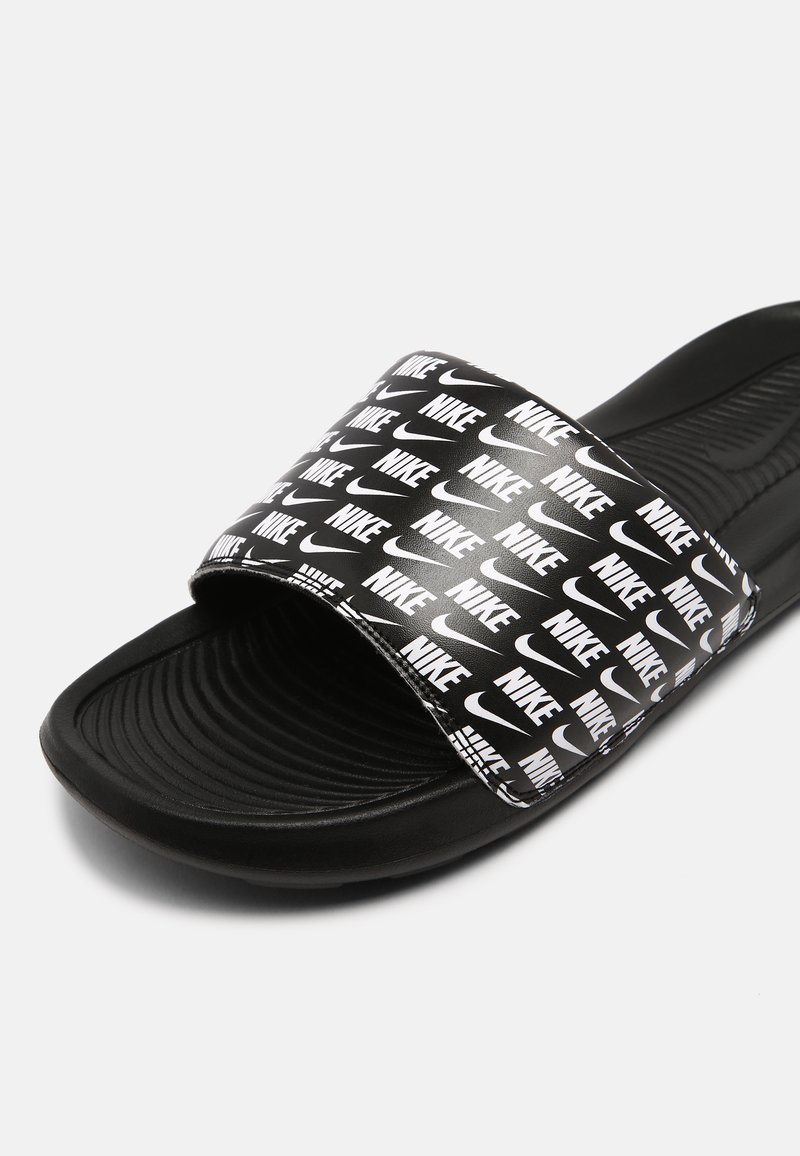 Chancla negra con suela texturizada, que presenta una banda ancha adornada con repetidos logotipos en blanco de "Nike". Material sintético suave.