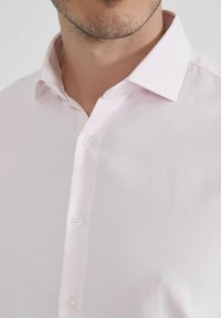 Chemise rose clair à col, en tissu texturé, avec des boutons blancs et un col pointu, révélant une partie du cou et de la poitrine.