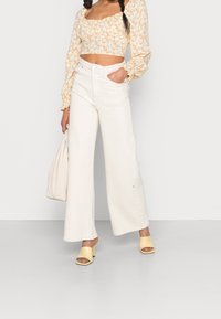 Vita vida byxor med hög midja, kombinerat med en blommig crop top med puffärmar och fyrkantig halsringning. Tillbehören inkluderar en beige väska och gula klackskor.