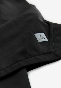 Chemise en tissu noir avec une texture lisse, présentant un patch logo triangulaire en gris sur la manche et des coutures détaillées le long de l'ourlet.
