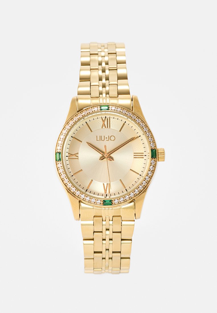 Goudkleurig horloge met een ronde wijzerplaat, een gestructureerde gouden schijf, Romeinse cijfers als markeringen en een met kristallen bezette bezel. Heeft groene accenten.