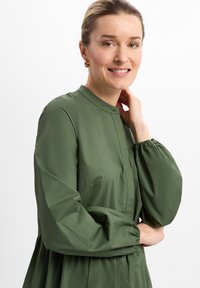 Grünes, leichtes Kleid mit hohem Kragen und langen, elastischen Ärmeln. Es hat eine geraffte Taille und einen subtilen Glanz in der Stofftextur.