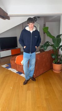 Νέος άνδρας με μπλε φούτερ με κουκούλα και τζιν στέκεται σε σαλόνι με καφέ καναπέ, φυτευτό φυτό, τηλεόραση, πορτοκαλί κουβέρτα και ξύλινο πάτωμα.