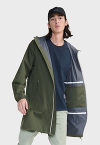 Veste de pluie imperméable de couleur vert olive avec un intérieur gris, fermeture éclair intégrale, poignets élastiques, poches latérales et accents réfléchissants.