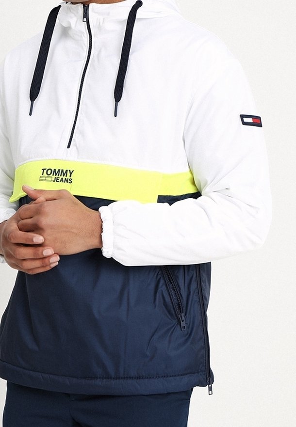 Vit och marinblå huoodad zip-jacka med en framsida dragkedja, gul horisontell rand och "TOMMY JEANS"-logotyp. Har elastiska ärmslut och sidofickor.