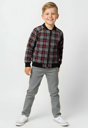 Młody chłopiec uśmiechający się, ubrany w czarno-czerwoną kratowaną kurtkę, szare spodnie i białe trampki, stojący na tle jednolitego jasnego tła.