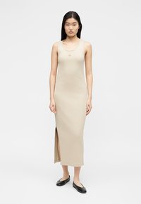 Νεαρή γυναίκα που φοράει μπεζ αμάνικο μάξι φόρεμα με πλαϊνή σχισμή και μικρή κεντητή λεπτομέρεια, συνοδευόμενο από μαύρα μπαλαρίνα παπούτσια, στέκεται ουδέτερα.