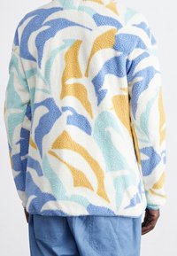 Veste en polaire avec une base crème ornée de motifs abstraits en vagues bleues, jaunes et turquoise. Fini texturé et doux. Ourlet à double couture.