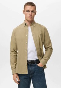 Camicia button-up color khaki chiaro con colletto a punta, maniche lunghe e vestibilità slim, indossata sopra una t-shirt bianca e abbinata a jeans scuri.