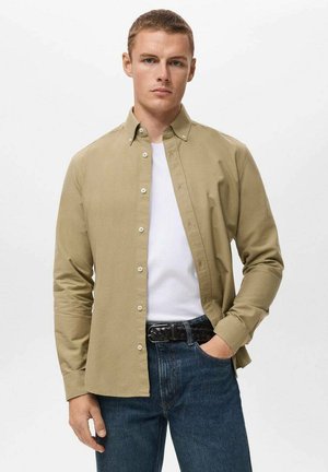 Light khaki button-up shirt with a pointed collar, long sleeves, and a slim fit, worn over a white t-shirt and paired with dark jeans.