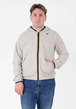Giacca cappuccio beige chiaro con zip frontale e una striscia colorata al centro. Caratterizzata da una tasca e polsini elasticizzati. Tessuto liscio e leggero.