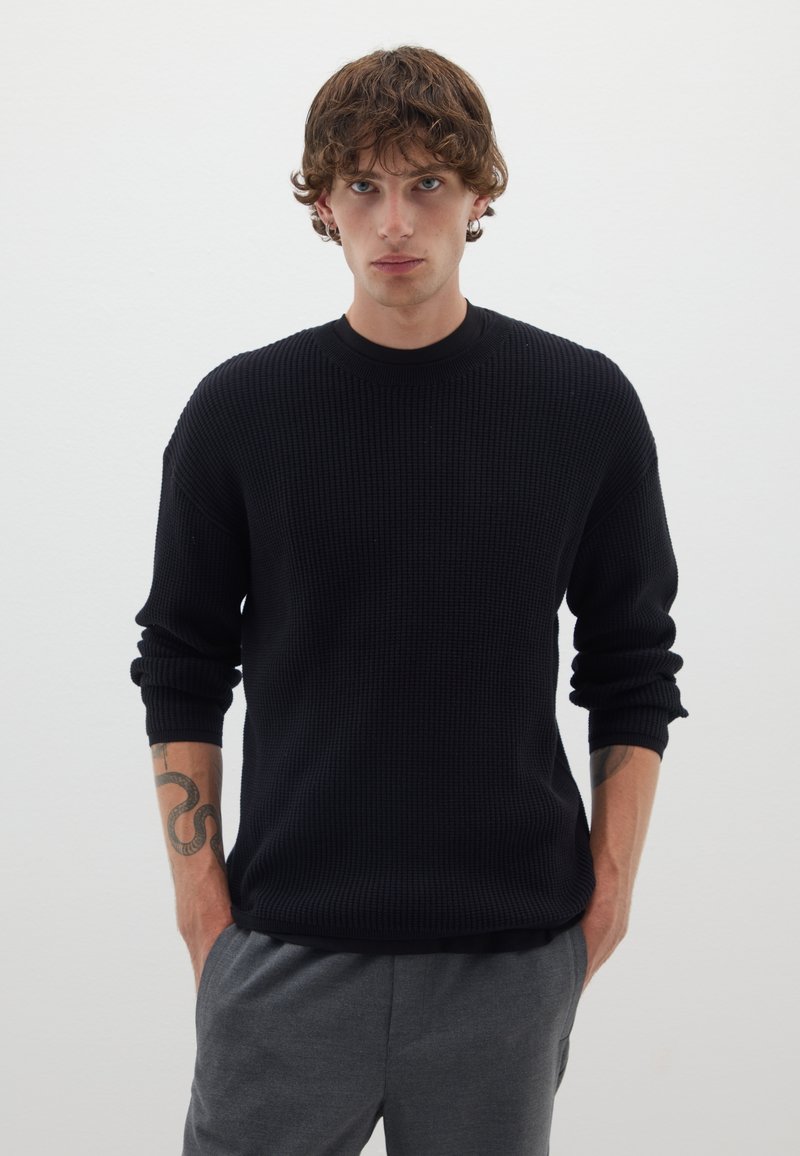 Pull en maille côtelée noir à manches longues, col rond et coupe décontractée. Associé à un pantalon gris avec cordon de serrage.