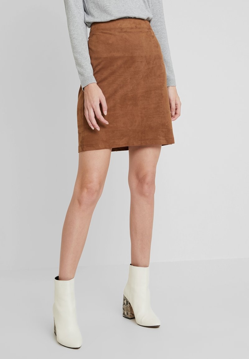 Femme portant une jupe en daim marron longueur genou, un haut à manches longues gris et des bottines blanches à talons à motifs, debout sur un sol gris.