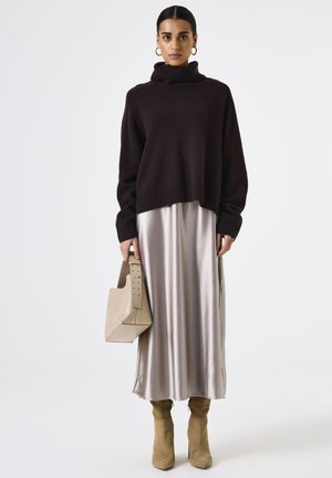 Bruine oversized coltrui, zilveren satijnen midi rok, beige gestructureerde handtas, tan kniehoge laarzen, model staand tegen een witte achtergrond.