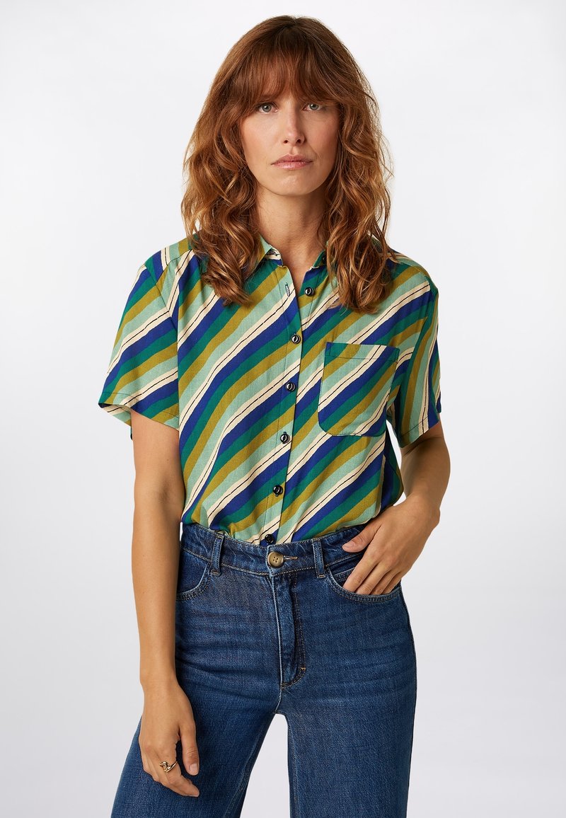 Short-sleeve shirt in green with blue and cream diagonal stripes. Features a front pocket and buttons, paired with dark blue jeans.