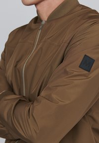 Brown bomber jacket with a smooth, shiny texture; features a ribbed collar, zip closure, and a black label on the left sleeve.