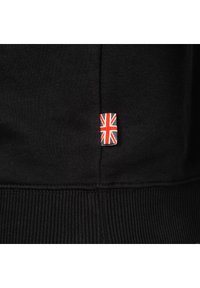 Svart sweatshirt med ribbad nederkant, som har en liten vävd etikett med den brittiska flaggan. Mjuk bomullstextur.