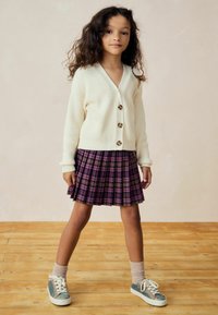Weiße, gerippte Strickjacke mit großen Knöpfen, kombiniert mit einem lila karotierten, gefältelten Rock. Funkelnde grüne Sneakers und hellrosa Socken vervollständigen das Outfit.
