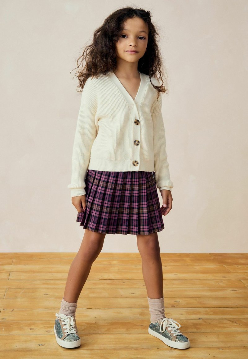 Weiße, gerippte Strickjacke mit großen Knöpfen, kombiniert mit einem lila karotierten, gefältelten Rock. Funkelnde grüne Sneakers und hellrosa Socken vervollständigen das Outfit.