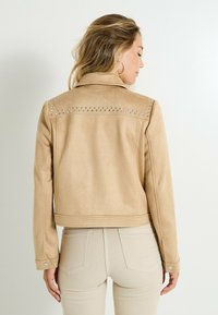 Veste cropped en daim beige avec des accents cloutés sur les épaules et le col ; présente des poignets à fermoir à pression et une texture lisse.