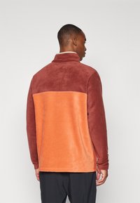 Fleece-pullover med hög krage, med en tvåtonad design i djup rödbrun och orange, mjuk textur och avslappnad passform.