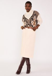 Veste en maille crème et marron avec des motifs géométriques, portée sur un col roulé blanc, associée à une jupe beige et des bottes à talons hauts marron.