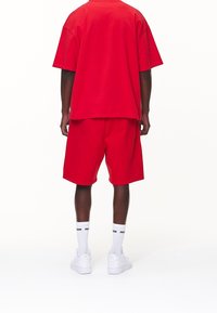 Červené bavlněné oversized tričko a odpovídající šortky, oboje v uvolněném střihu. Bílá tenisky a ponožky dotvářejí celý outfit.