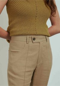 Pantalon en mélange de lin brun clair avec poches avant et fermeture par bouton, associé à un débardeur en maille moutarde texturée.