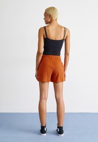 Svart ärmlös topp med tunna axelremmar, kombinerad med rostfärgade plisserade shorts och svarta högskor, bild tagen bakifrån.