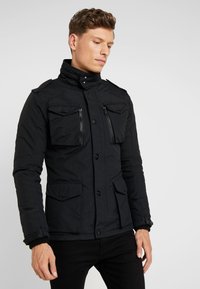Black padded jacket featuring a high collar, multiple front pockets, and zippered chest pockets. Textured fabric with a matte finish.