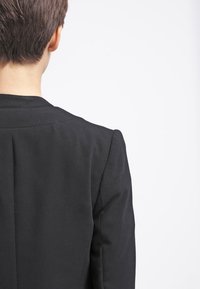 Černé strukturované sako s přiléhavým střihem, čistými ramenními švy a hladkým textilním povrchem. Zadní pohled zdůrazňuje elegantní linie a jednoduchost.
