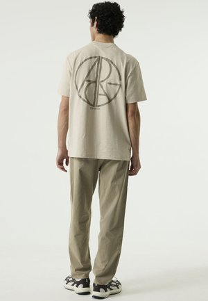 Man staat met de rug naar de camera, draagt een beige t-shirt met een groot rond grafisch ontwerp, lichte broek en zwart-witte sneakers.