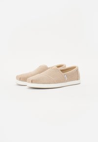 Zapatillas beige tipo slip-on con tejido texturizado, que presentan rayas horizontales, puntera redonda y suela blanca. Logo sutil en el lateral.